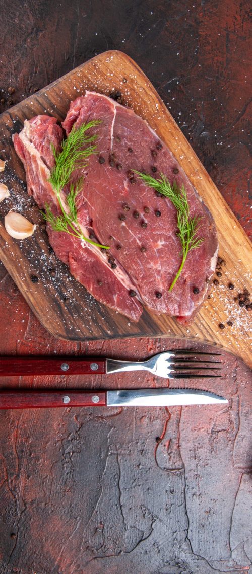 Overhead view of red meat on wooden cutting board and garlic green pepper fork and knife on dark background