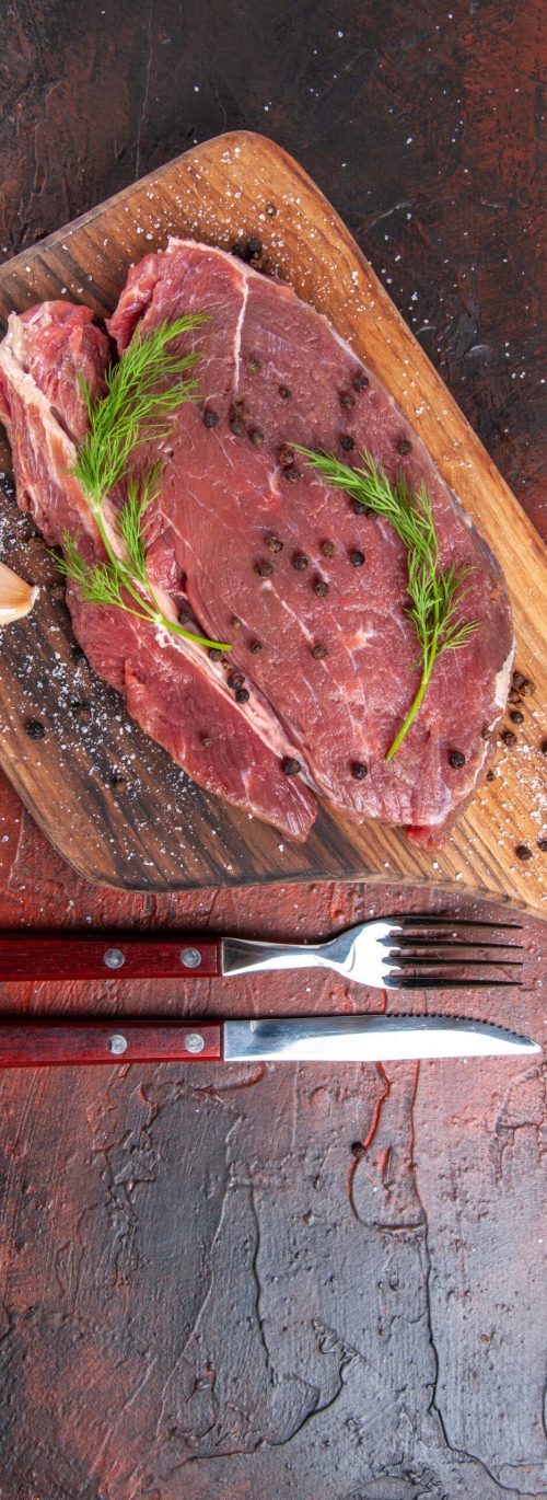 Overhead view of red meat on wooden cutting board and garlic green pepper fork and knife on dark background
